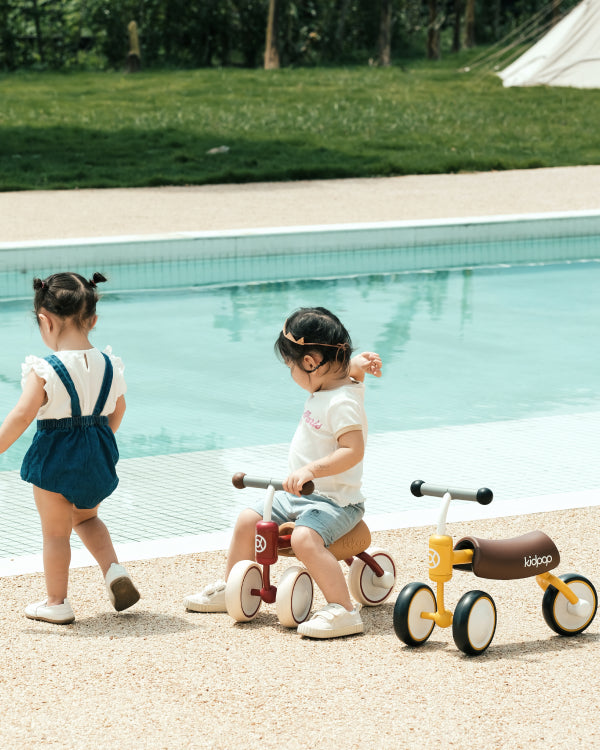 Toddlers riding red and yellow Kidpop BEE Baby Balance Bikes poolside.