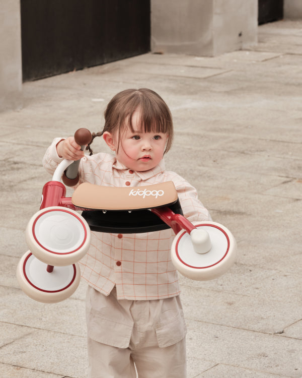 Toddler with red and tan BEE Baby Balance Bike, 'kidpop' logo on seat, designed for learning balance.