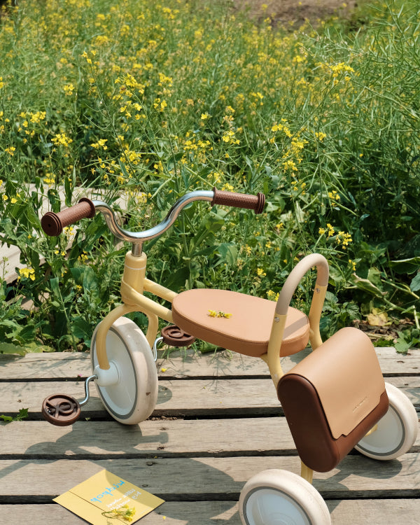 MATE Kids' yellow tricycle with brown seat, handlebars, pedals, and saddle bag, on wooden deck with wildflowers.