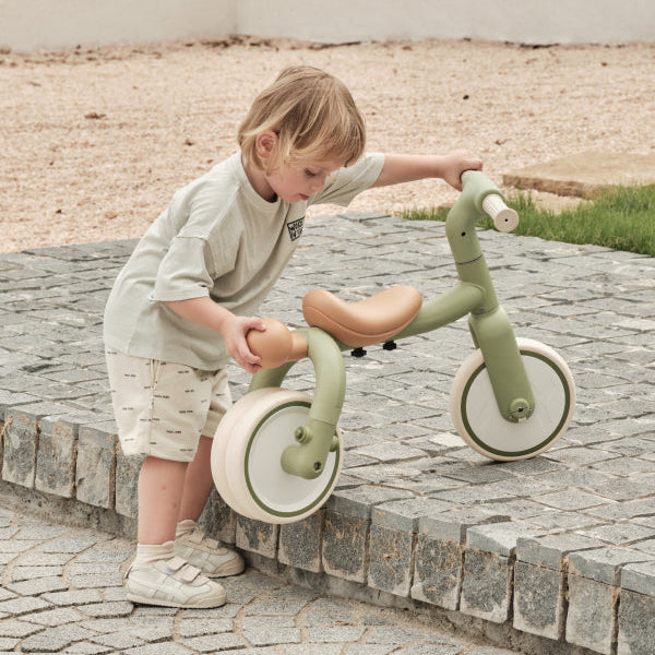 Green and tan PONY 4-in-1 kids' tricycle with tan seat, a young boy interacts with it on a stone path.