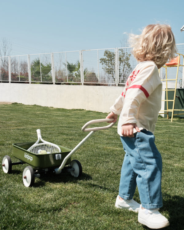 Child pulling olive green PULA-Wild Lite Kid's Wagon with tennis racket and balls on lawn.
