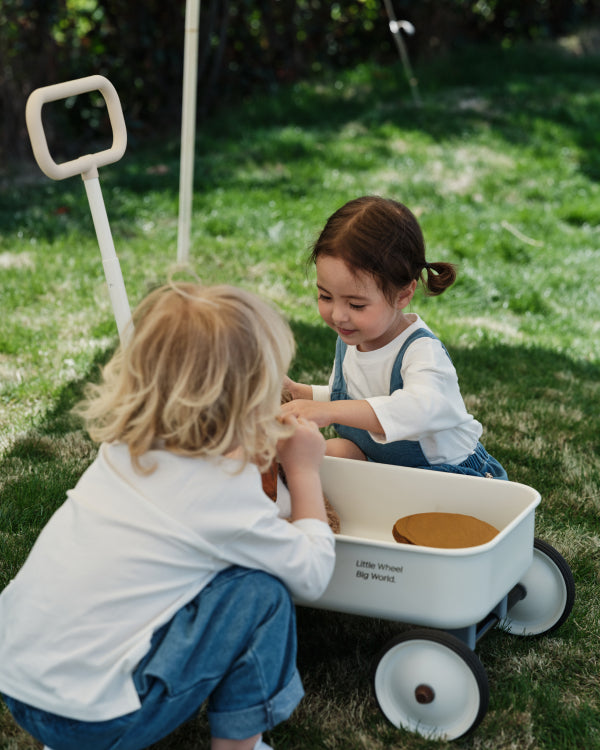Two children playing with a cream PULA-Wild Lite Kid's Wagon on grass. Wagon features 'Little Wheel Big World' text.