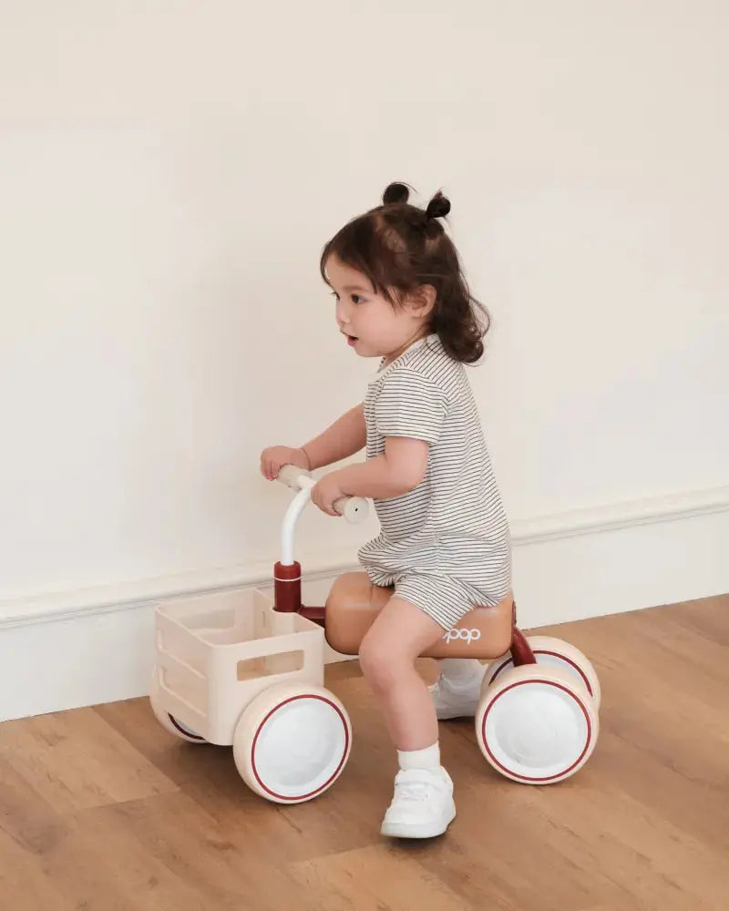 Child riding TOAST Baby Balance Bike with storage basket and red accents on wooden floor.