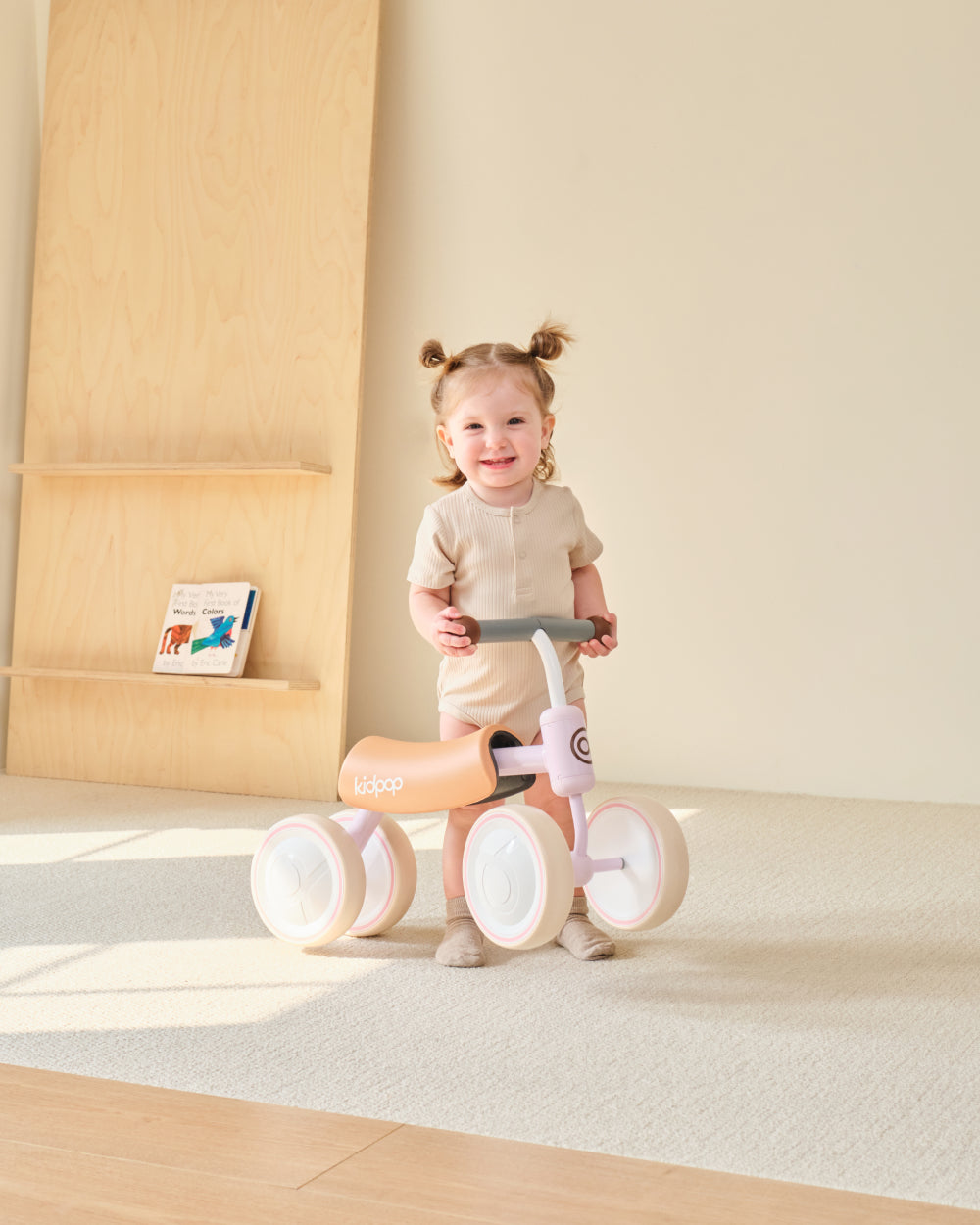 Kidpop BIG BEE pink and tan four-wheel balance bike with happy toddler girl indoors.