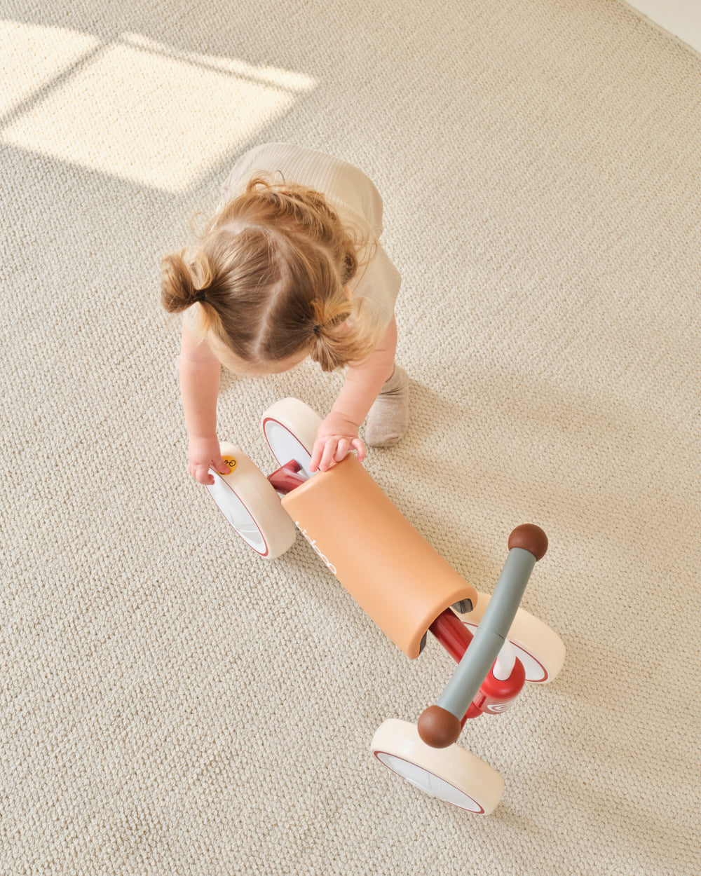 BIG BEE Baby Balance Bike, child plays with tan seat, cream wheels, gray handlebar indoors.