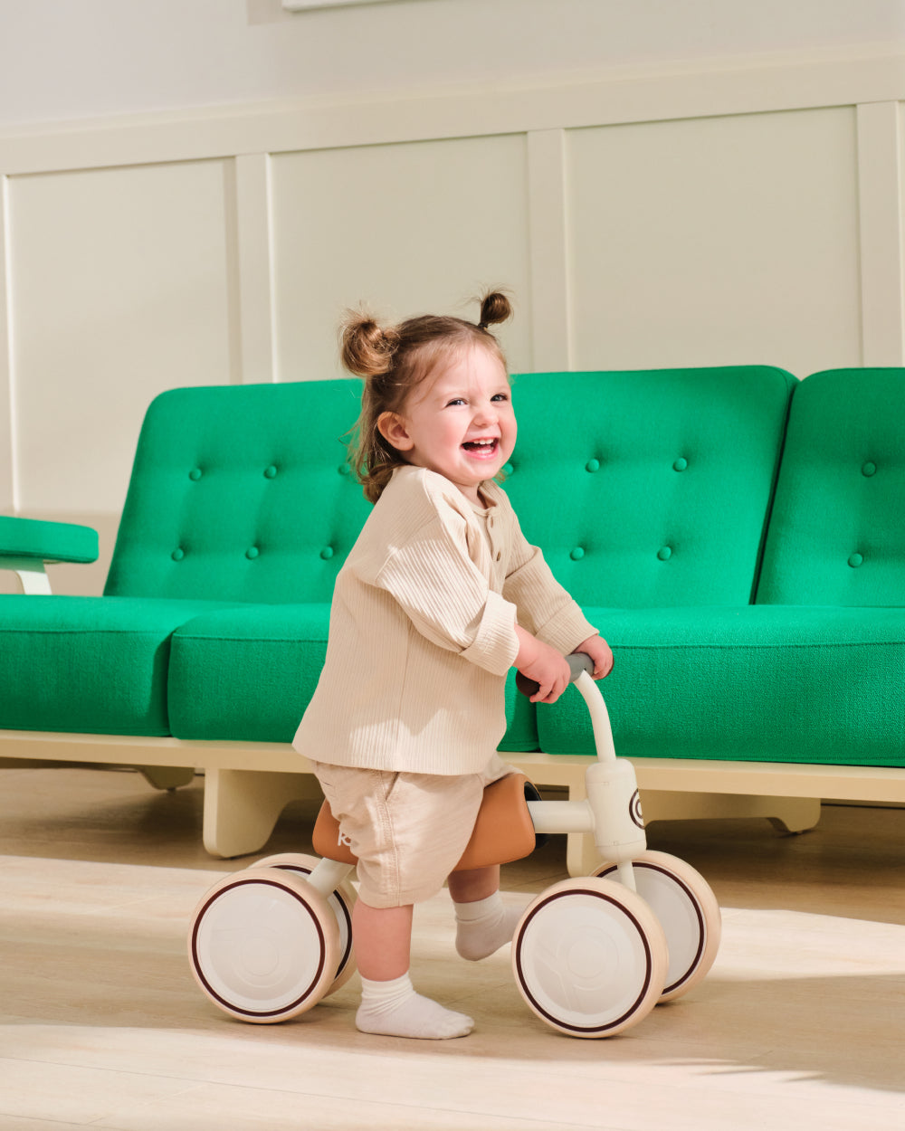 Toddler happily riding a BIG BEE white and tan four-wheel balance bike indoors, featuring cream wheels and tan seat.