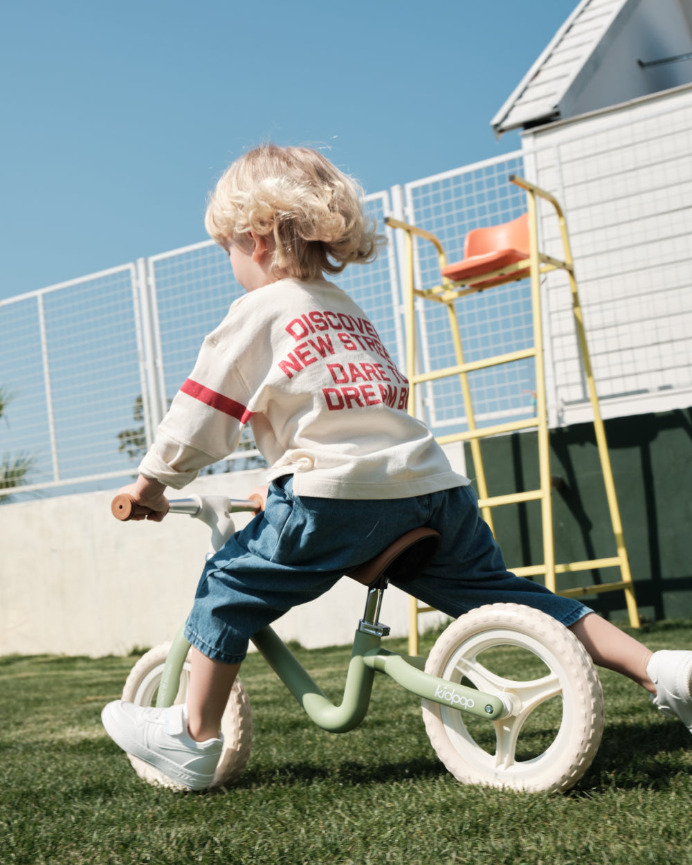 Child riding sage green RIGHT balance bike with brown seat and tan grips, cream wheels, outdoor fun on product page.