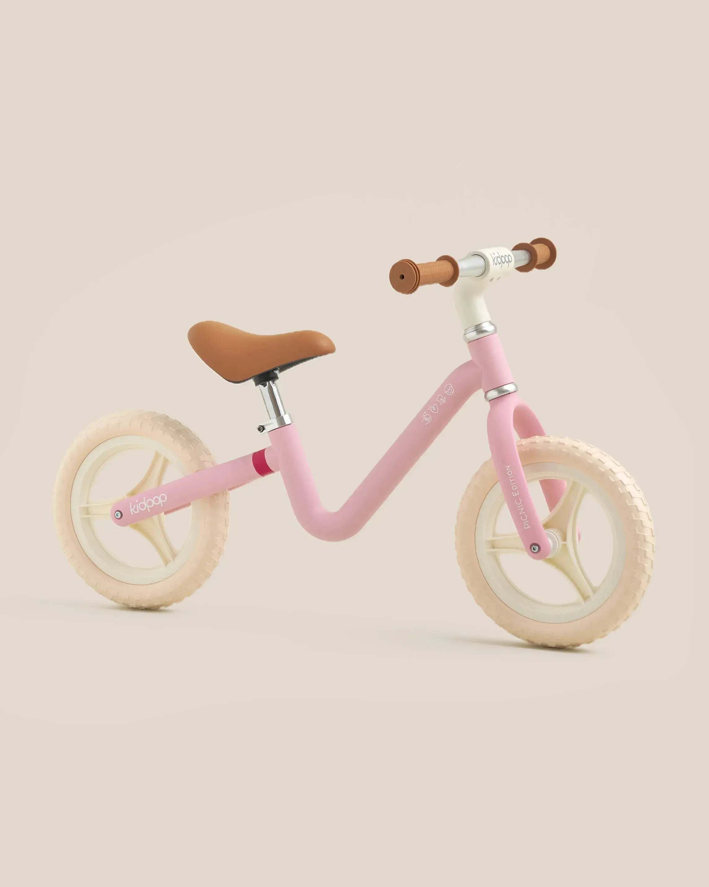 Pale pink RIGHT balance bike with brown seat and tan handlebars, cream wheels, showcasing minimalist design on product page.