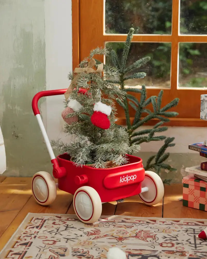 Red Kidpop PULA wagon with Christmas tree and miniature stockings, featuring cream wheels and white handle, on patterned rug.