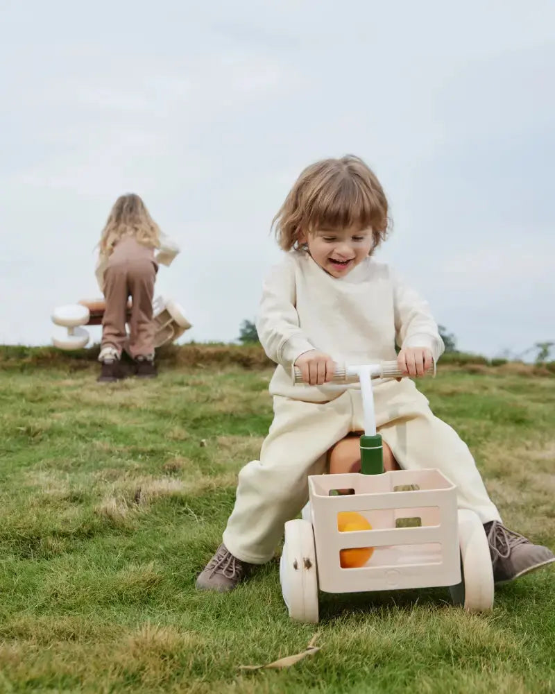 Smiling child riding beige TOAST baby balance bike with green accents and storage basket in grassy field.