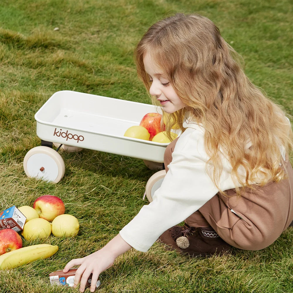 A young girl playing with a toy wagon and fruit on the grass.