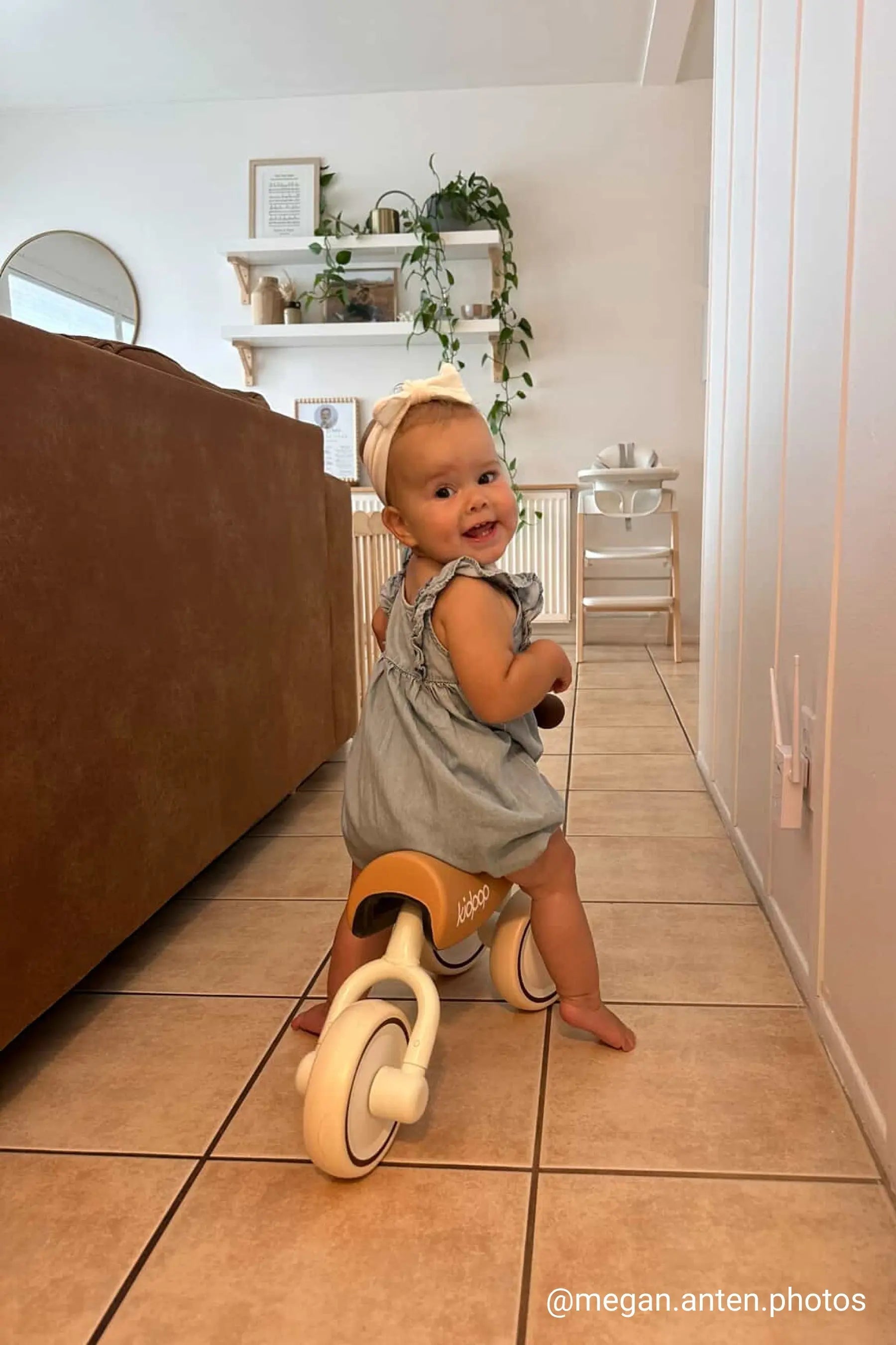 A smiling toddler in a blue dress riding a wooden balance bike indoors.