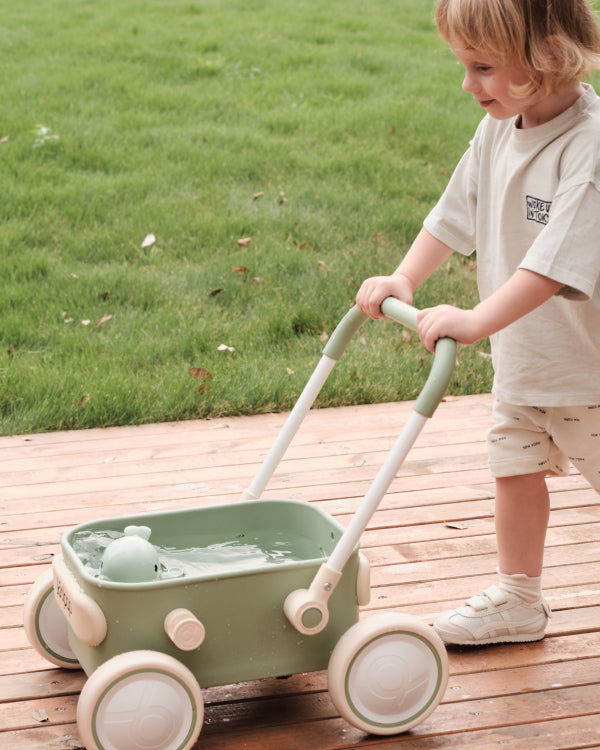 Little child pushing a green Kidpop wagon filled with water and a toy on a wooden deck.