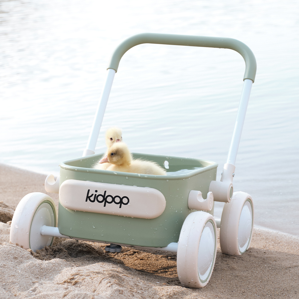 Two yellow ducklings floating in a sage green Kidpop toy wagon filled with water on a sandy beach.
