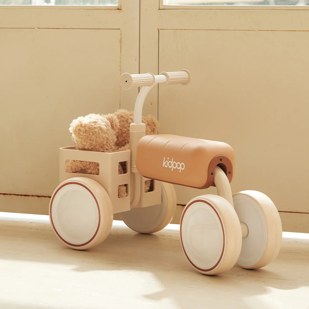 A tan Kidpop balance bike with a teddy bear sitting in the front basket.