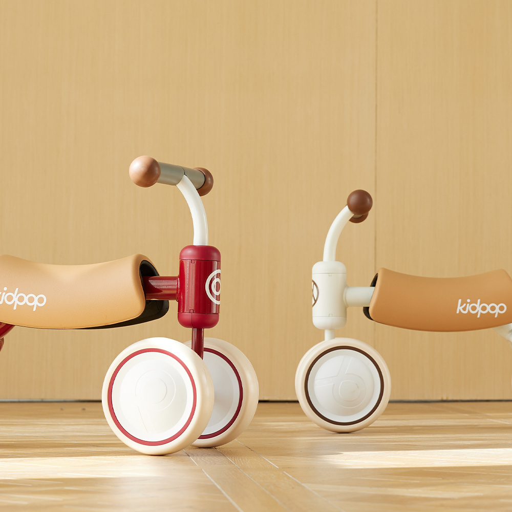 A close-up shot of two Kidpop toddler balance bikes, one red and one white, standing on a sunlit wooden floor.