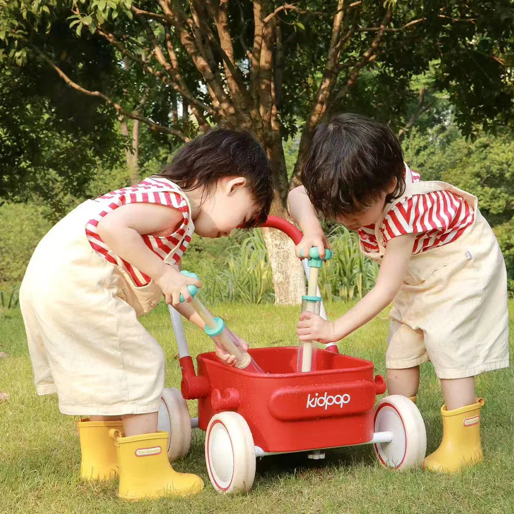 Two toddlers playing with toy pumps in a red Kidpop wagon on a grassy field.