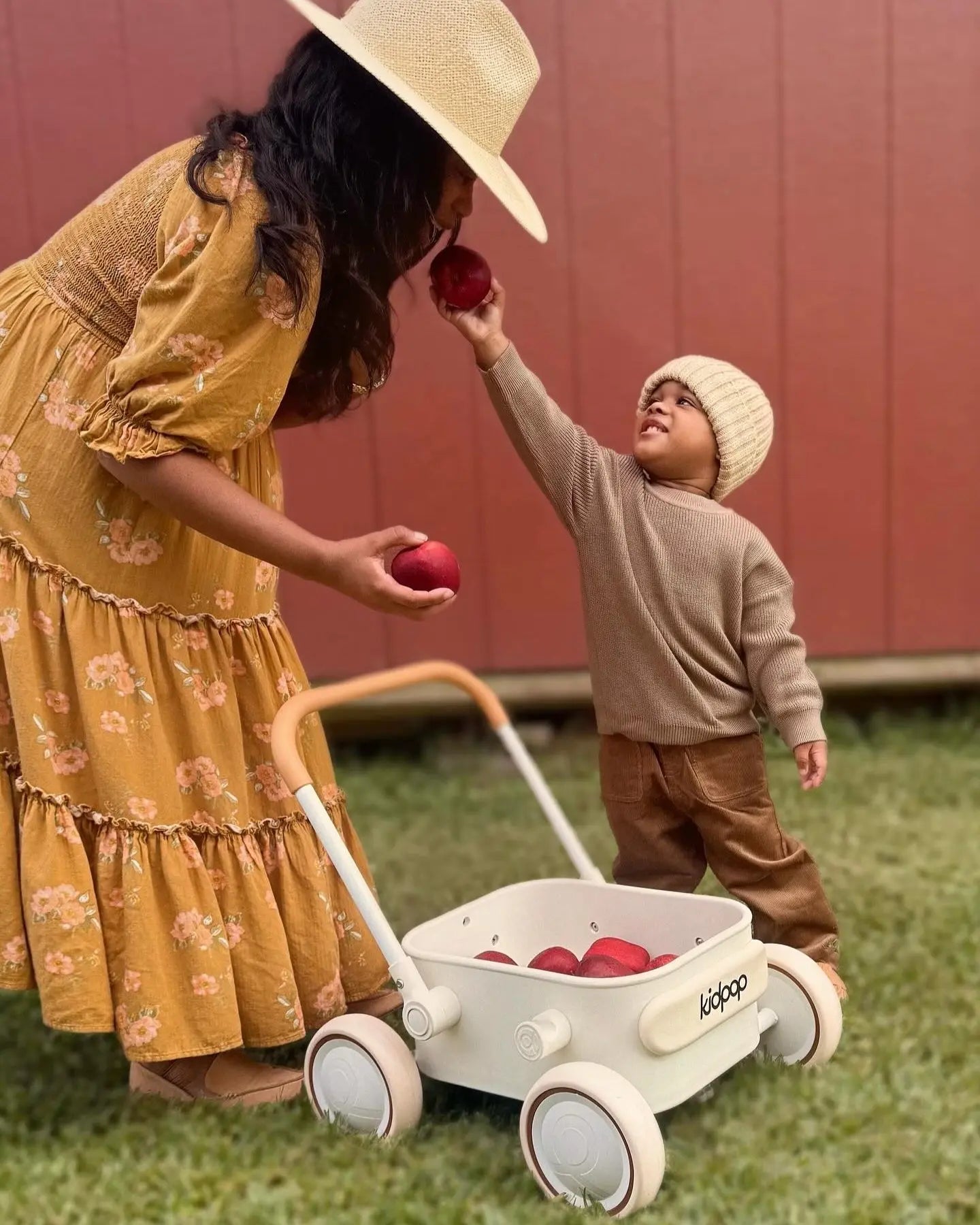 Woman in a yellow dress and straw hat interacting with a child near a white toy cart outdoors.